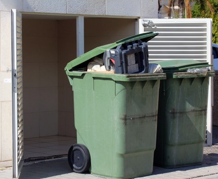 A green plastic refuse bin with a partially open lid, revealing a white sack and some scattered rubbish inside, positioned on a concrete driveway adjacent to a small, open-fronted storage shed with a tiled floor and a light-colored interior wall. The bin is located near a white slatted fence or panel, and the background includes a glimpse of trees or greenery indicating an outdoor residential area. The scene suggests a typical waste collection point for a property in Cricklewood, with the bin ready for rubbish removal services. The bin's surface shows some dirt and marks, consistent with regular outdoor use. The lighting is natural, casting soft shadows on the pavement and highlighting the texture of the plastic and the surrounding environment. Commercial Waste Cricklewood offers rubbish removal services, as indicated by the context of the scene, which emphasizes waste management in a local residential setting.