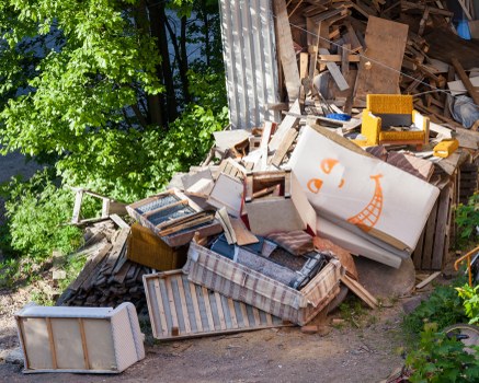 A large pile of discarded household furniture and wooden debris situated outdoors beside a corrugated metal structure, with a backdrop of green leafy trees. The pile includes a striped upholstered armchair, a white wooden wardrobe or cabinet, a mattress or large cushion with a checked cover, and various wooden planks and panels of differing sizes, textures, and finishes. Some items appear weathered and cluttered, with parts leaning or stacked unpredictably. A yellow plastic tool box and other smaller objects are visible amongst the debris. The scene is illuminated by natural daylight, highlighting the contrast between the materials and the lush greenery surrounding the rubbish. This image illustrates the type of waste that Commercial Waste Cricklewood's rubbish removal service in Cricklewood might handle for residential or commercial clients in the area.