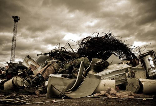 A large pile of scrap metal and discarded appliances situated outdoors on a dirt or asphalt surface, with visible rust, dents, and peeling paint on the metallic objects, including washing machines, refrigerators, and industrial components. The debris is haphazardly arranged, with some items leaning or partially overlapping others, and a tangled mass of wire or metal at the top of the pile. In the background, there is a tall metal tower structure against a cloudy, overcast sky, suggesting an industrial or waste disposal site, possibly within or near Cricklewood. The overall scene is captured with a muted, monochrome tone, emphasizing the texture and deterioration of the waste materials, consistent with the context of rubbish removal services offered by Commercial Waste Cricklewood.