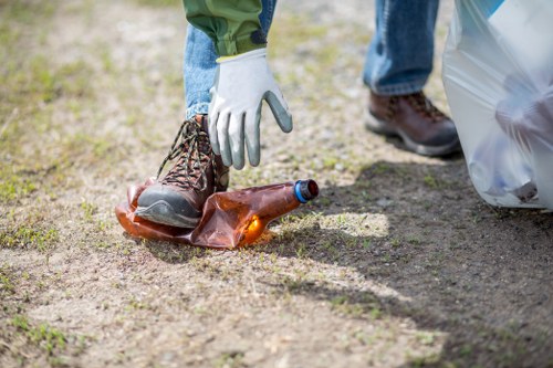 Close-up image of a person wearing white gloves and brown work boots, reaching down to pick up a crumpled brown glass bottle from a patchy outdoor ground surface that appears to be a mixture of soil and sparse grass. In the background, a clear plastic rubbish bag is partially visible on the right side, nearby a pair of blue jeans and darker shoes, suggesting a rubbish removal or cleanup activity. The environment is typical of an outdoor area where waste collection or litter clearance might take place, with natural daylight illuminating the scene. This image highlights the process of rubbish gathering, relevant to services like commercial waste removal in Cricklewood, and demonstrates the careful handling and collection of glass waste for proper disposal or recycling, consistent with standards required by companies such as Commercial Waste Cricklewood serving the local postcode area.