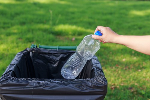 A person's hand is seen holding a clear plastic water bottle by the neck, about to place it into a black plastic rubbish bin lined with a black liner. The bin is situated outdoors on a paved or concrete surface, with a background of well-maintained green grass and natural daylight enhancing the scene. The water bottle is partially crushed, transparent, with a blue cap at the top. The environment suggests a typical waste collection area or garden setting, reflecting proper disposal of plastic waste. The image emphasizes the importance of rubbish management and environmentally responsible waste disposal, consistent with services offered by Commercial Waste Cricklewood in the Cricklewood area. The detailed focus on the water bottle and disposal bin highlights household or commercial rubbish collection activities, supporting local SEO relevance related to waste removal services in the postcode region. The overall tone is neutral and observational, with emphasis on the practical aspect of rubbish disposal.