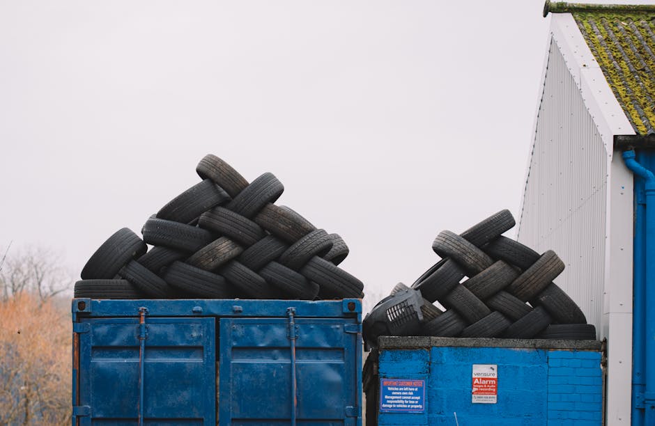 A large blue metal skip filled with stacked used black rubber tires, positioned outdoors next to a white building with a partially moss-covered roof. The tires, arranged in an overlapping pattern, var