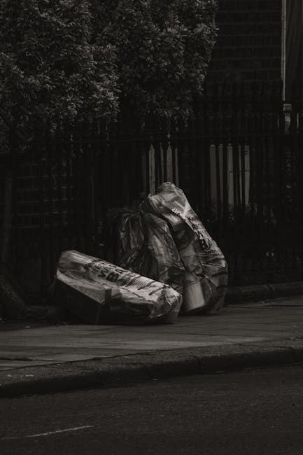 A group of large black rubbish bags filled with waste are placed on a sidewalk close to a black metal fence, with some leaning against it. The bags appear to be made of thick plastic, with visible cre