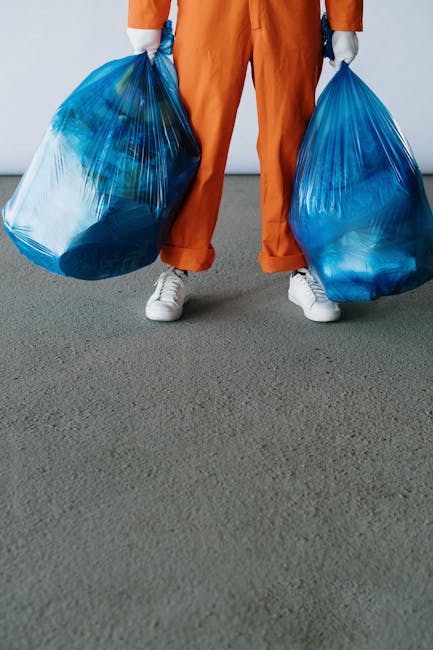 A group of large black rubbish bags filled with waste are placed on a sidewalk close to a black metal fence, with some leaning against it. The bags appear to be made of thick plastic, with visible cre