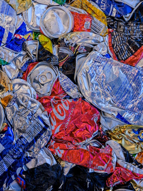 A close-up view of a pile of crumpled aluminum cans and crushed plastic beverage bottles, primarily silver with some red, black, and blue branding visible. The cans display various textures, from smoo